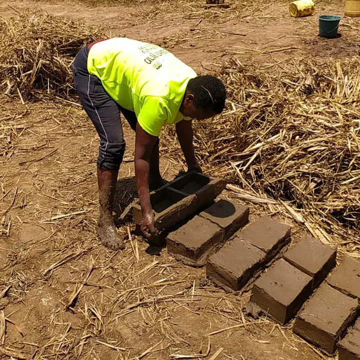 A woman molds wet clay into bricks on a sunny day as part of a Destiny Impact CBO initiative supporting women’s economic empowerment through sustainable brickmaking.
